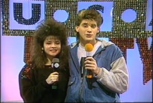 Two Dance Party USA cast members on camera in front of the studio sign