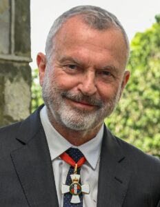 Sam Neill smiling in a dark suit and wearing his New Zealand knighthood insignia on a red ribbon in 2022