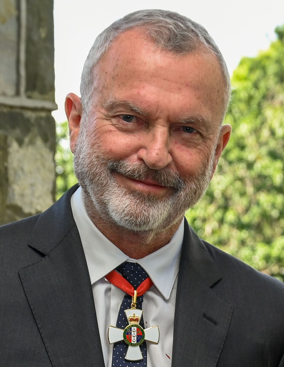 Sam Neill smiling in a dark suit and wearing his New Zealand knighthood insignia on a red ribbon in 2022