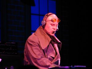 Thomas Dolby performing live behind a bank of keyboards in goggles and a headset microphone