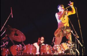 Phil Collins singing at the microphone while Chester Thompson plays drums behind him, Genesis on stage in 1981